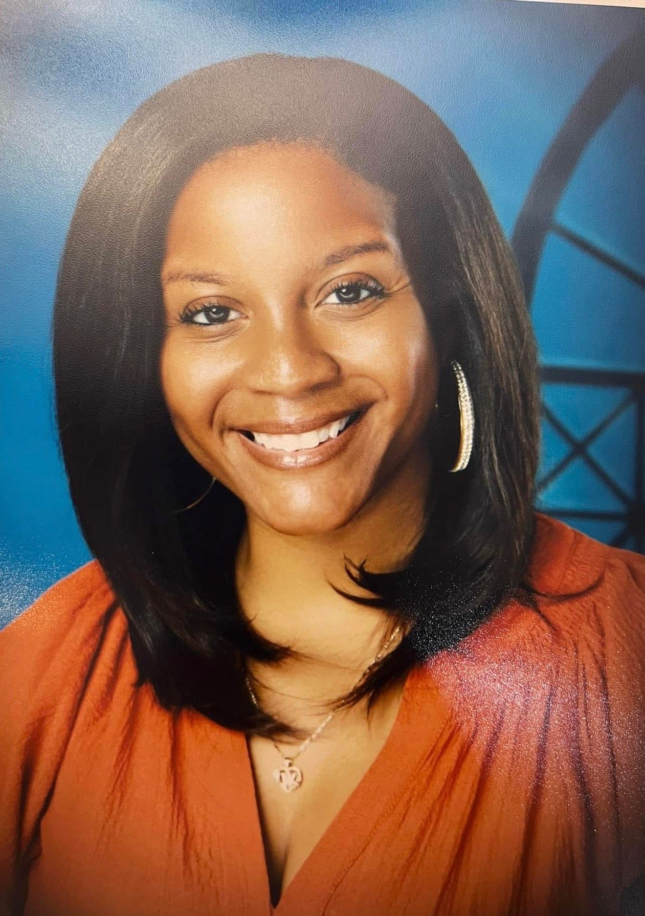 Smiling woman with long hair wearing an orange top and earrings against a blue background.